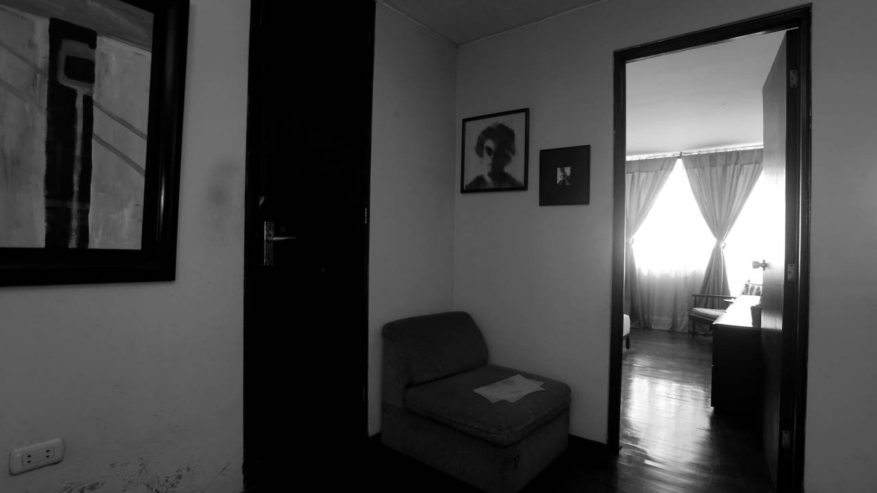 Czarno-biała fotografia pozioma, kompozycja otwarta. Wnetrze jasnego mieszkania z meblami w stylu retro. W lewym górnym rogu widoczny minimalistyczny obraz w masywnej ramie. Dalej widoczne czarne drzwi, obok mały fotel przy ścianie. Nad fotelem zawieszone dwa obrazki. Po prawej stronie wejście do naturalnie, jasno oświetlonego pomieszczenia z oknami wyposażonymi w długie firany i związane zasłony.